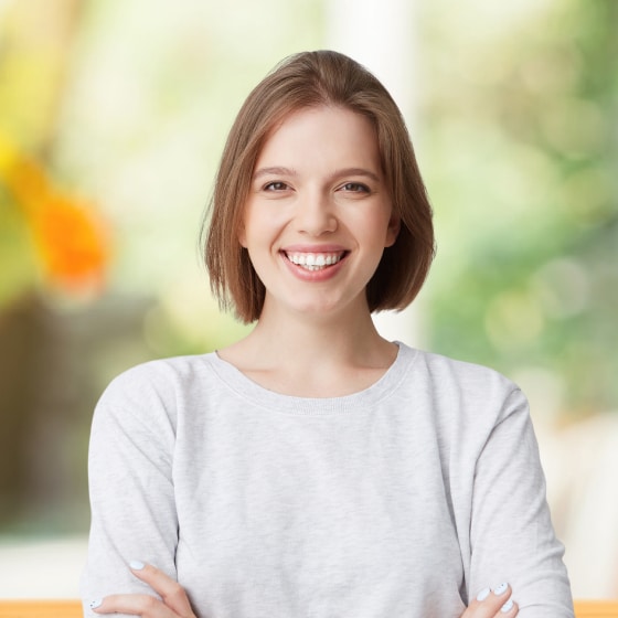 A person with short brown hair is smiling and crossing their arms, wearing a light gray sweater. The background is softly blurred with green and orange hues.
